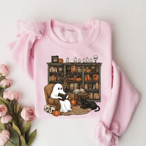 May include: A pink sweatshirt with a graphic of a ghost reading a book in a library with a black cat and pumpkins. The graphic is in a cartoon style.