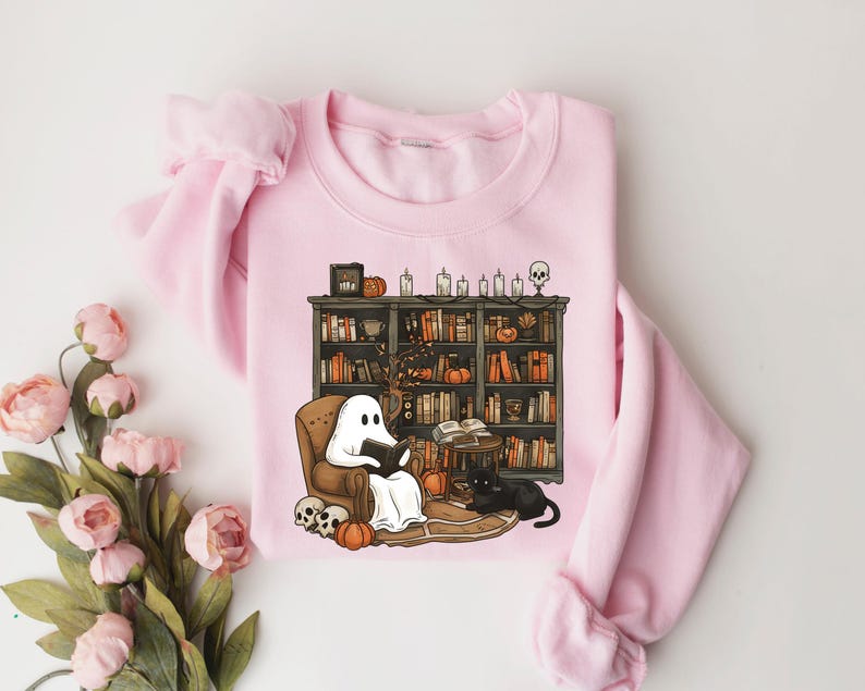May include: A pink sweatshirt with a graphic of a ghost reading a book in a library with a black cat and pumpkins. The graphic is in a cartoon style.