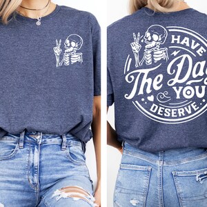May include: A navy blue t-shirt with a white skeleton graphic on the front and a white circle graphic on the back that says "Have The Day You Deserve".
