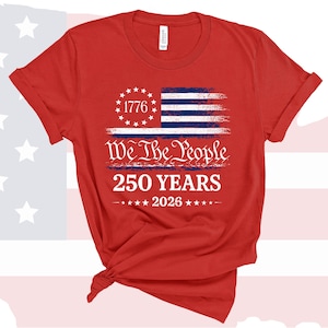 May include: A red short-sleeved t-shirt featuring a graphic of a distressed American flag with a circle of stars and the number 1776. Below this, the text reads We The People, 250 YEARS, and 2026, accented by a row of small white stars.