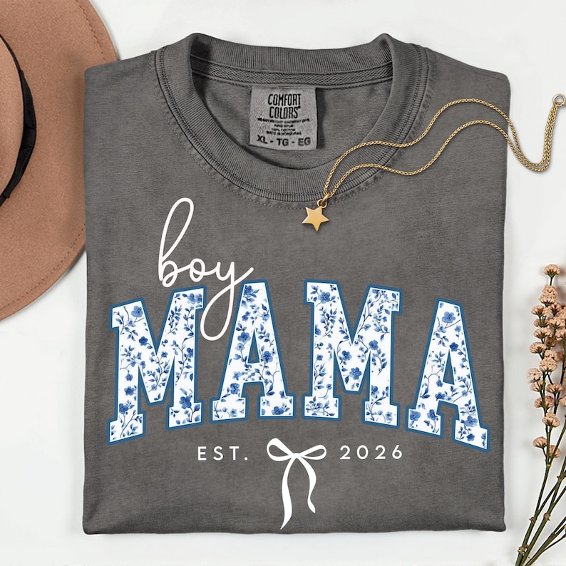 以下が含まれることがあります： 白文字と青い花柄で「boy MAMA」と書かれたグレーのXL Comfort Colors Tシャツ。 シャツには「EST. 2026」とリボンのグラフィックが含まれています。 金色の星のペンダントネックレスと茶色の帽子も表示されています。