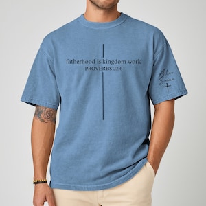 May include: A blue t-shirt with the text "fatherhood is kingdom work PROVERBS 22:6" and a vertical line design. The shirt also features the signature "Alex Susan" with a cross symbol.