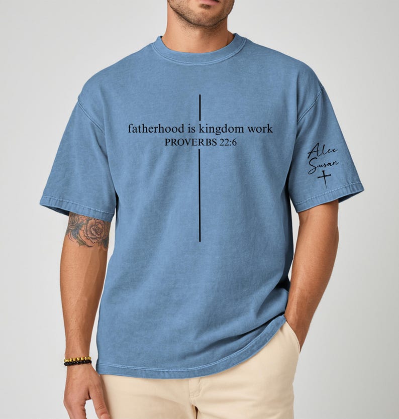 May include: A blue t-shirt with the text "fatherhood is kingdom work PROVERBS 22:6" and a vertical line design. The shirt also features the signature "Alex Susan" with a cross symbol.