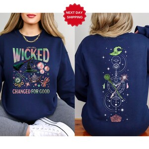 Puede incluir: Sudadera azul marino con la palabra "WICKED" en una fuente colorida y degradada. La parte delantera presenta un sombrero de bruja, una escoba y otros elementos m&aacute;gicos. La parte trasera tiene un sombrero de bruja, una escoba y un dise&ntilde;o celestial. El texto "CHANGED FOR GOOD" tambi&eacute;n est&aacute; en la parte delantera.