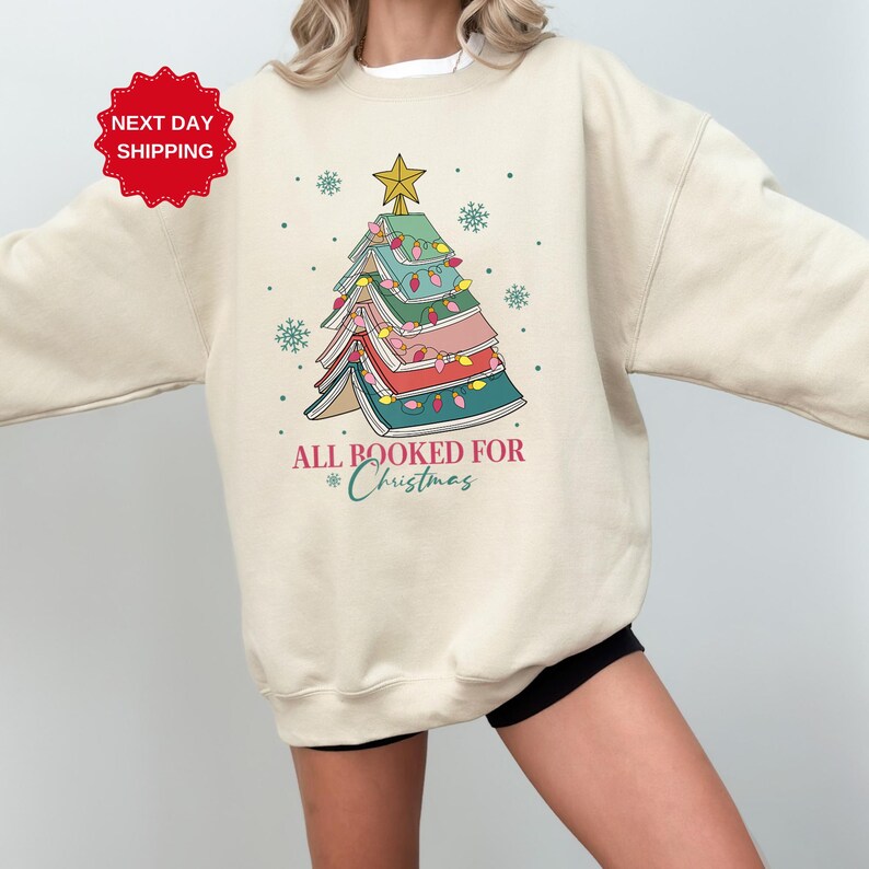 Puede incluir: Una sudadera de color crema con un dise&ntilde;o de &aacute;rbol de Navidad hecho de libros apilados. El &aacute;rbol est&aacute; decorado con luces de colores y una estrella en la parte superior. El texto "All Booked for Christmas" est&aacute; impreso debajo del &aacute;rbol.