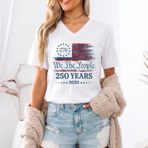 May include: A white V-neck t-shirt featuring a distressed red and blue American flag graphic with a circle of stars and the text 1776. Below the flag, the text reads We The People, 250 YEARS, and 2026 with small stars on each side.
