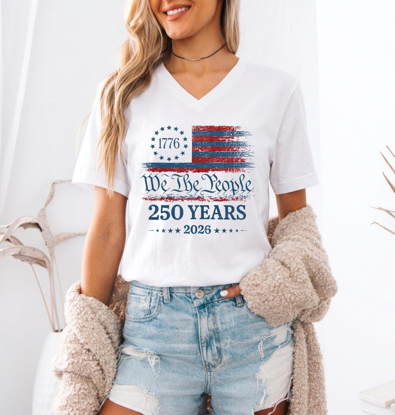 May include: A white V-neck t-shirt featuring a distressed red and blue American flag graphic with a circle of stars and the text 1776. Below the flag, the text reads We The People, 250 YEARS, and 2026 with small stars on each side.