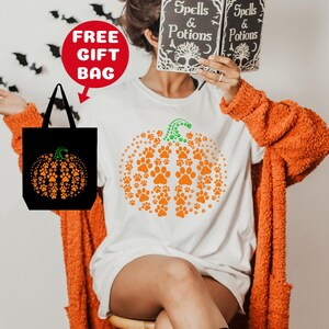 May include: White t-shirt with an orange pumpkin design made of paw prints. The pumpkin has a green stem. The t-shirt is being modeled by a person wearing a fuzzy orange cardigan. A black tote bag with a similar pumpkin design is included.