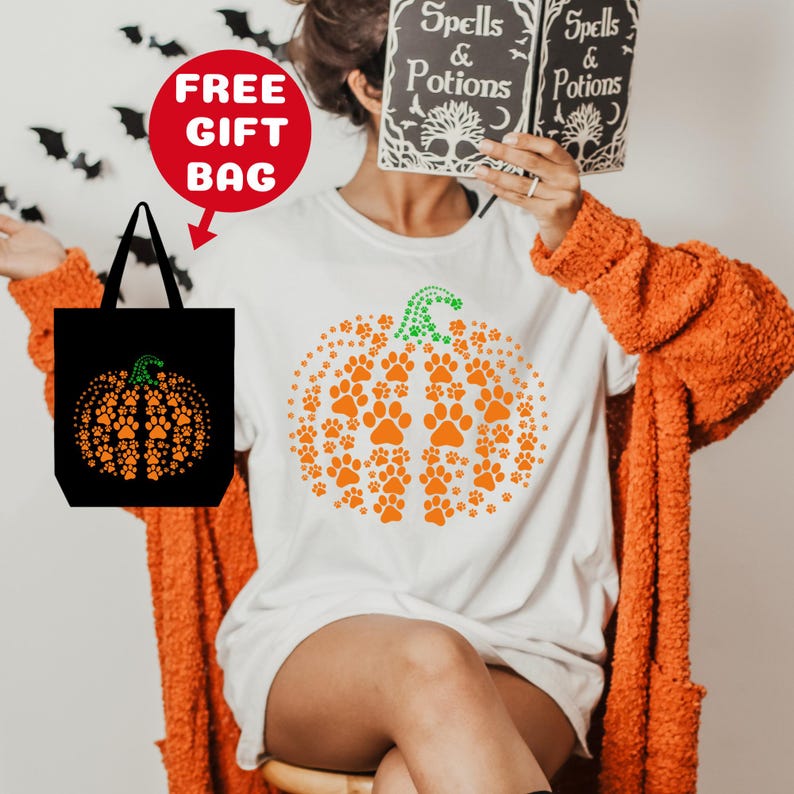 May include: White t-shirt with an orange pumpkin design made of paw prints. The pumpkin has a green stem. The t-shirt is being modeled by a person wearing a fuzzy orange cardigan. A black tote bag with a similar pumpkin design is included.