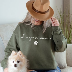 May include: A woman wearing a green sweatshirt with the text "dog mom" and a white paw print. She is holding a small, fluffy, white dog.