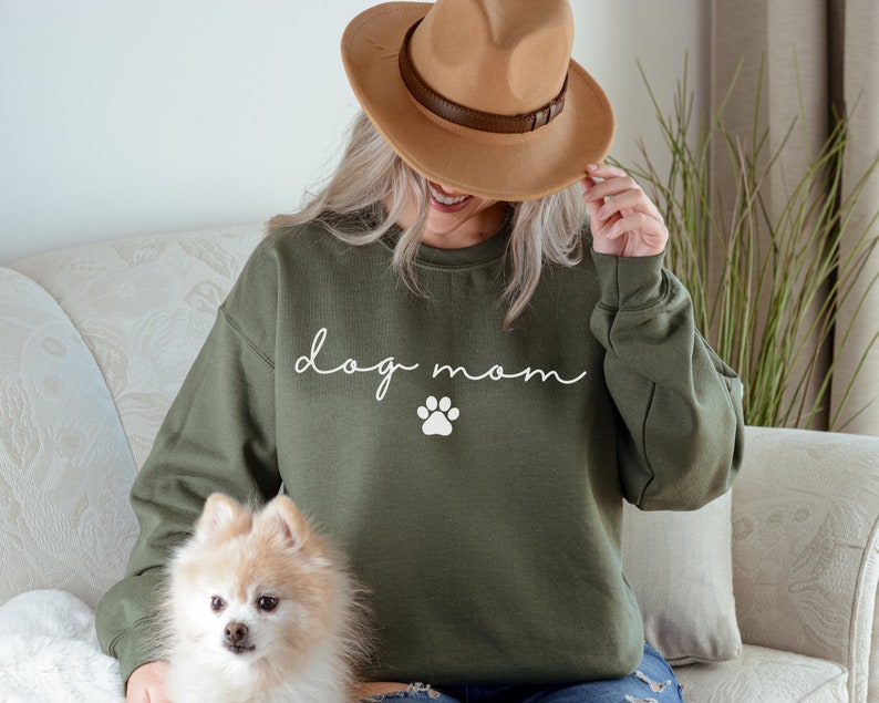 May include: A woman wearing a green sweatshirt with the text "dog mom" and a white paw print. She is holding a small, fluffy, white dog.