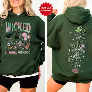 Puede incluir: Sudadera con capucha verde oscuro con el texto "WICKED CHANGED FOR GOOD" en una fuente colorida y caprichosa. La parte delantera presenta un sombrero de bruja, una escoba y burbujas. La parte trasera tiene un dise&ntilde;o a juego con un sombrero y elementos celestiales.