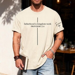 May include: Cream-colored t-shirt with the text "fatherhood is kingdom work PROVERBS 22:6" and a vertical black line forming a cross. The sleeve has the name "Susan" and a cross. The shirt is a casual, oversized fit.