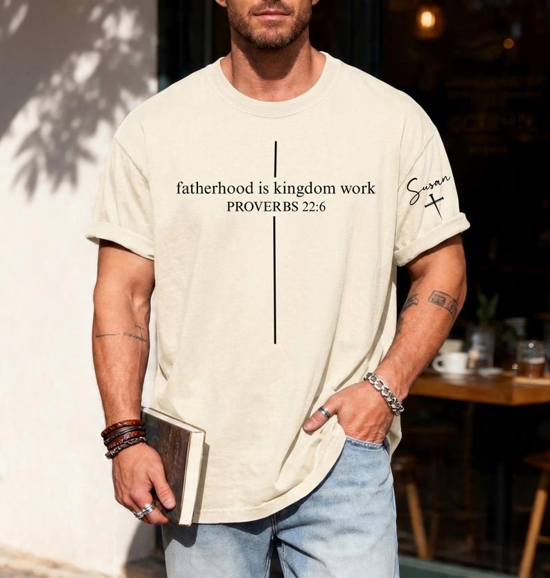May include: Cream-colored t-shirt with the text "fatherhood is kingdom work PROVERBS 22:6" and a vertical black line forming a cross. The sleeve has the name "Susan" and a cross. The shirt is a casual, oversized fit.