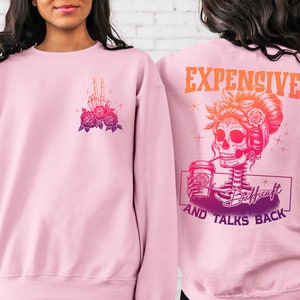 May include: Pink sweatshirt with a graphic of a skeleton woman with a coffee cup and the text "Expensive and Talks Back". The skeleton woman is wearing a flower crown and has a skull face. The sweatshirt has a pink to orange ombre effect.