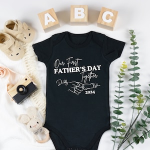 May include: Black baby onesie with white text that reads "Our First Father's Day Together" with a hand holding a smaller hand graphic and the year 2024. The text "Daddy" and "[Baby's Name]" are written in cursive.