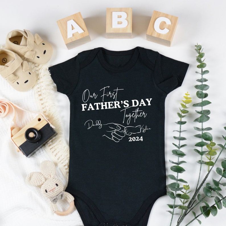 May include: Black baby onesie with white text that reads "Our First Father's Day Together" with a hand holding a smaller hand graphic and the year 2024. The text "Daddy" and "[Baby's Name]" are written in cursive.