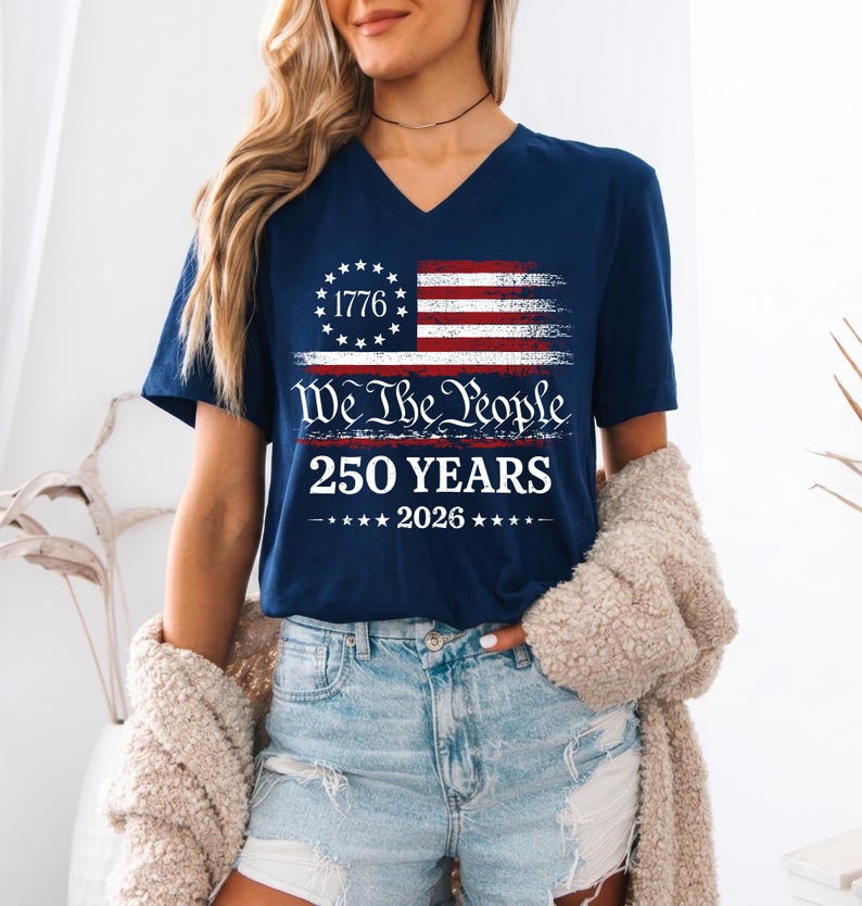 May include: A navy blue V-neck t-shirt featuring a graphic of a distressed American flag with a circle of stars and the text 1776. Below the flag, the text reads We The People, 250 YEARS, and 2026, accented by small stars on each side of the year.