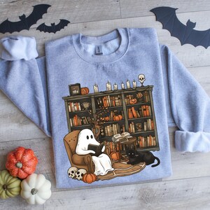 May include: A gray sweatshirt with a graphic of a ghost reading a book in a cozy living room with a bookshelf full of books, a black cat, and pumpkins. The ghost is wearing a white sheet and has a friendly smile.