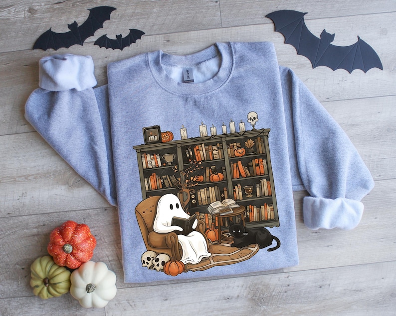 May include: A gray sweatshirt with a graphic of a ghost reading a book in a cozy living room with a bookshelf full of books, a black cat, and pumpkins. The ghost is wearing a white sheet and has a friendly smile.