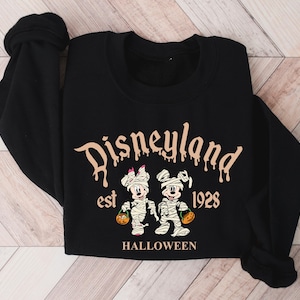 May include: Black sweatshirt with a white and brown graphic of Mickey Mouse and Minnie Mouse dressed as mummies. The text "Disneyland est 1928 HALLOWEEN" is printed on the sweatshirt.
