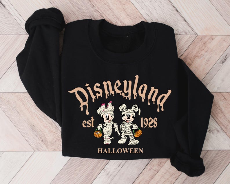 May include: Black sweatshirt with a white and brown graphic of Mickey Mouse and Minnie Mouse dressed as mummies. The text "Disneyland est 1928 HALLOWEEN" is printed on the sweatshirt.