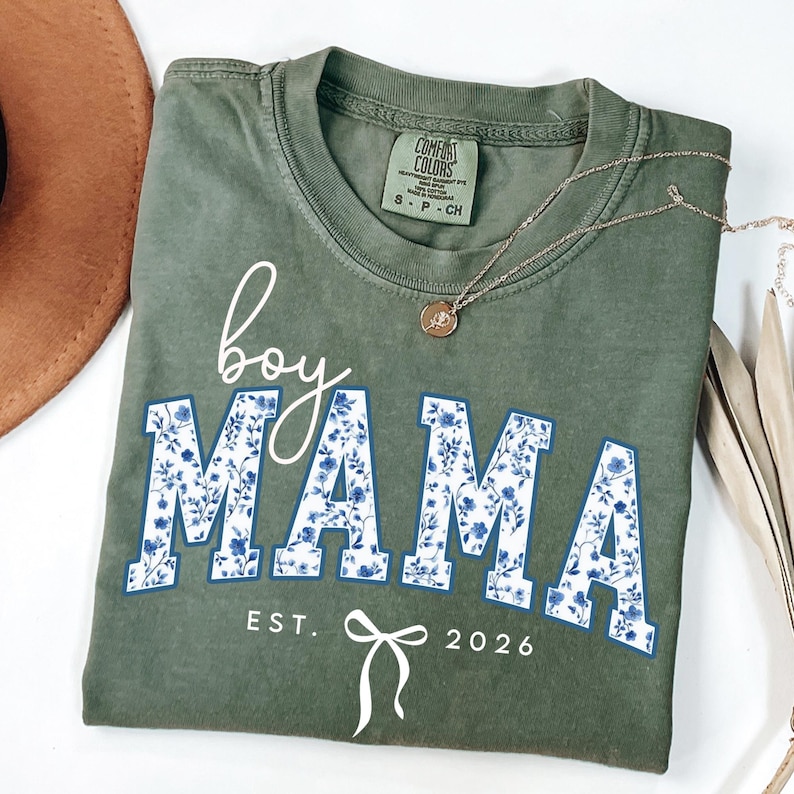 以下が含まれることがあります： オリーブグリーンのTシャツ。白の筆記体で「boy MAMA」と青い花柄がプリントされています。「EST. 2026」の文字とリボンのグラフィックが下にあります。ペンダント付きのゴールドネックレスがシャツの上にあります。