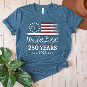 May include: A heather blue short-sleeve t-shirt featuring a distressed graphic of the American flag with a circle of stars and the year 1776. Below the flag, the text reads We The People, 250 YEARS, and 2026 with stars on either side of the year.
