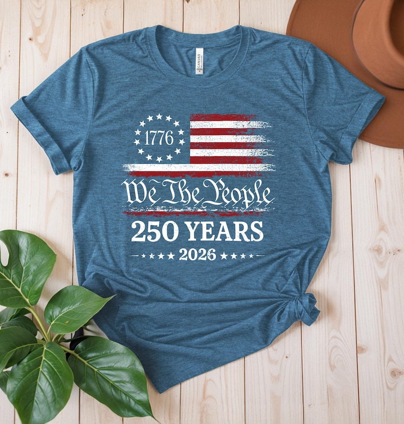 May include: A heather blue short-sleeve t-shirt featuring a distressed graphic of the American flag with a circle of stars and the year 1776. Below the flag, the text reads We The People, 250 YEARS, and 2026 with stars on either side of the year.