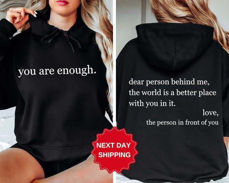 Puede incluir: Sudadera con capucha negra con el texto "you are enough." en la parte delantera y "dear person behind me, the world is a better place with you in it. love, the person in front of you" en la parte trasera.