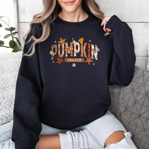 Może przedstawiać: Granatowa bluza z napisem "PUMPKIN SEASON" w jesiennym wzornictwie. Tekst zawiera mieszankę kolor&oacute;w pomarańczowego, brązowego i białego, z akcentami liści i gwiazd. Bluza to casualowy element garderoby na jesień.