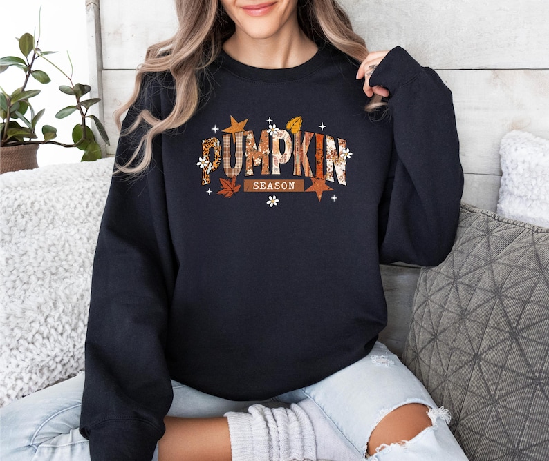 Może przedstawiać: Granatowa bluza z napisem "PUMPKIN SEASON" w jesiennym wzornictwie. Tekst zawiera mieszankę kolor&oacute;w pomarańczowego, brązowego i białego, z akcentami liści i gwiazd. Bluza to casualowy element garderoby na jesień.
