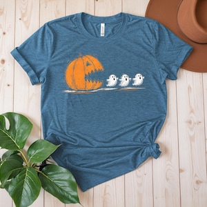 May include: A blue t-shirt featuring a Halloween-themed graphic. The design shows an orange pumpkin with a jagged mouth chasing three white ghosts. The shirt is made of a soft material and has a classic crew neck.