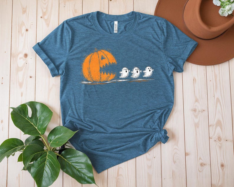 May include: A blue t-shirt featuring a Halloween-themed graphic. The design shows an orange pumpkin with a jagged mouth chasing three white ghosts. The shirt is made of a soft material and has a classic crew neck.
