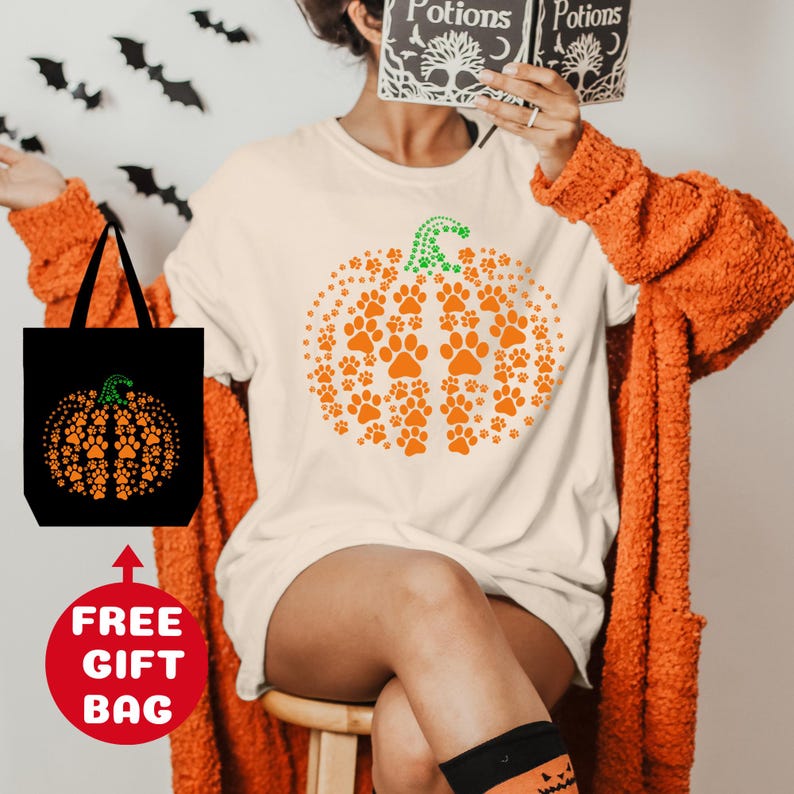 May include: A white t-shirt with an orange pumpkin design made of paw prints. The pumpkin has a green stem and is surrounded by smaller paw prints. The t-shirt is being modeled by a person wearing a long-sleeved orange cardigan. The image also includes a black tote bag with a similar pumpkin design.