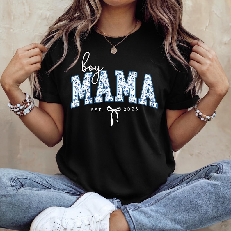 以下が含まれることがあります： 青い花柄の「boy MAMA」の文字が入った黒いTシャツ。「EST. 2026」とリボンのグラフィックも含まれています。Tシャツはクルーネックです。
