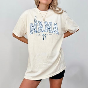 以下が含まれることがあります： クリーム色のTシャツ。青い花柄の文字で「boy MAMA」と書かれています。シャツには「EST. 2026」の文字と青いリボンのグラフィックも付いています。半袖でゆったりとしたフィット感です。