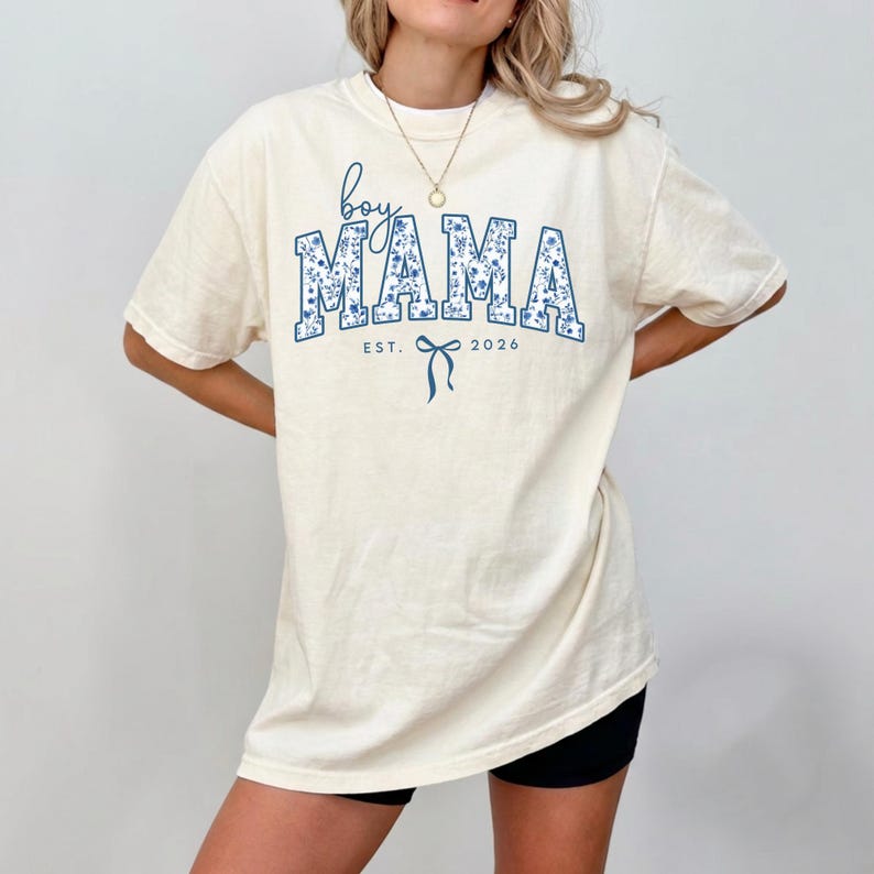 以下が含まれることがあります： クリーム色のTシャツ。青い花柄の文字で「boy MAMA」と書かれています。シャツには「EST. 2026」の文字と青いリボンのグラフィックも付いています。半袖でゆったりとしたフィット感です。