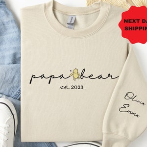 May include: A beige sweatshirt with the words "papa bear" in a script font, accompanied by a Winnie the Pooh graphic. Below the text is "est. 2023". The right sleeve has the names "Olivia" and "Emma" in a script font.