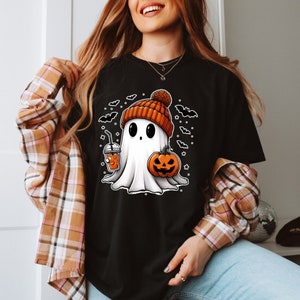 Może przedstawiać: Czarny T-shirt z uroczym kresk&oacute;wkowym duchem w pomarańczowo-brązowym kapeluszu, trzymającym dynię i nap&oacute;j. Duch jest otoczony przez nietoperze.