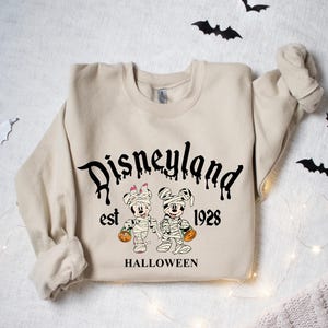 May include: A beige sweatshirt with a black graphic print of Mickey and Minnie Mouse dressed as mummies. The text "Disneyland est 1928 HALLOWEEN" is printed below the graphic.