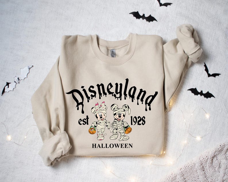 May include: A beige sweatshirt with a black graphic print of Mickey and Minnie Mouse dressed as mummies. The text "Disneyland est 1928 HALLOWEEN" is printed below the graphic.