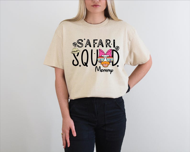 K&ouml;nnte beinhalten: Beiges T-Shirt mit dem schwarzen Aufdruck "SAFARI SQUAD Mommy", verziert mit einem Safarihut und Sonnenbrillen-Grafik. Das Shirt ist ein l&auml;ssiger Stil mit kurzen &Auml;rmeln.