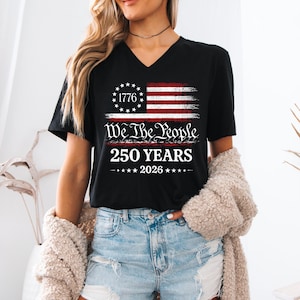 May include: A black V-neck t-shirt featuring a graphic of a distressed American flag with a circle of stars and the number 1776. Below the flag, the text reads We The People, 250 YEARS, and 2026, centered in white and red lettering on the chest.