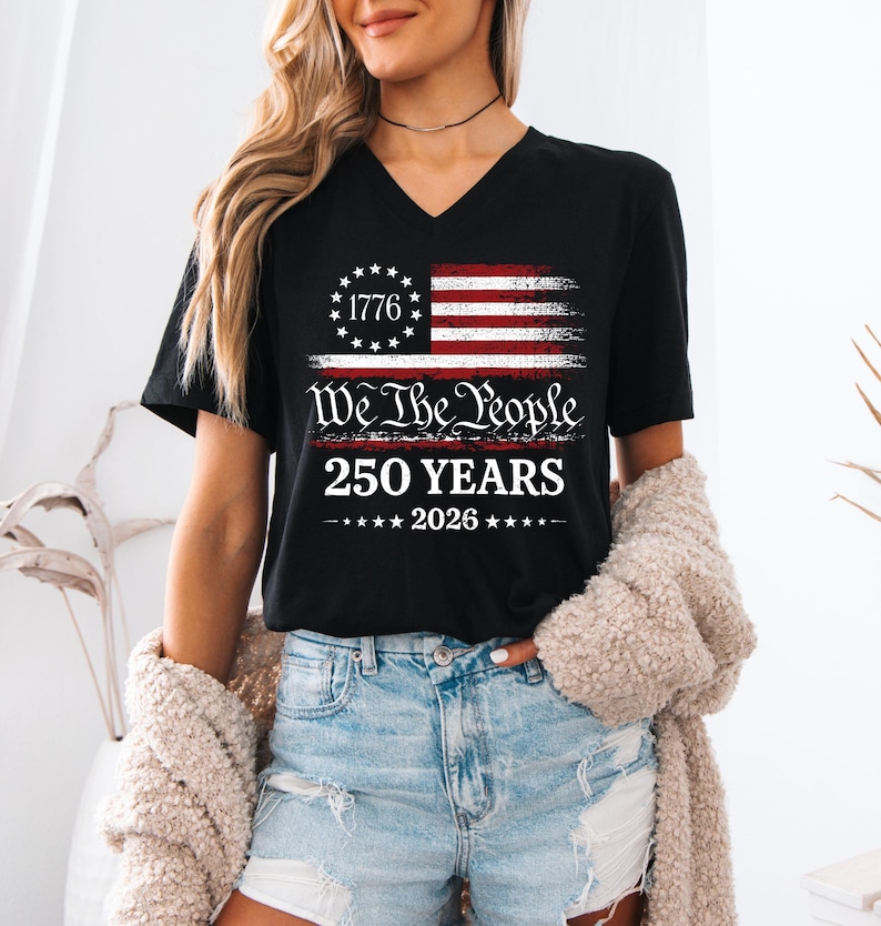 May include: A black V-neck t-shirt featuring a graphic of a distressed American flag with a circle of stars and the number 1776. Below the flag, the text reads We The People, 250 YEARS, and 2026, centered in white and red lettering on the chest.