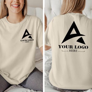 May include: A beige t-shirt with a black graphic of a stylized letter A with the text "YOUR LOGO HERE" on the front and back.
