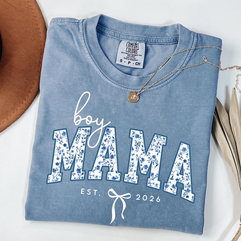 以下が含まれることがあります： 白の筆記体と花柄プリントで「boy MAMA」と書かれた青色のComfort Colors Tシャツ。テキスト「EST. 2026」とリボンが下にあります。ペンダント付きのゴールドネックレスがシャツの上にかけられています。