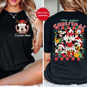 Op de afbeelding: Zwart T-shirt met een kerstthema met Mickey Mouse en andere Disney-figuren. Het shirt heeft de tekst "Prettige Kerst" en heeft een aanpasbaar naamveld.