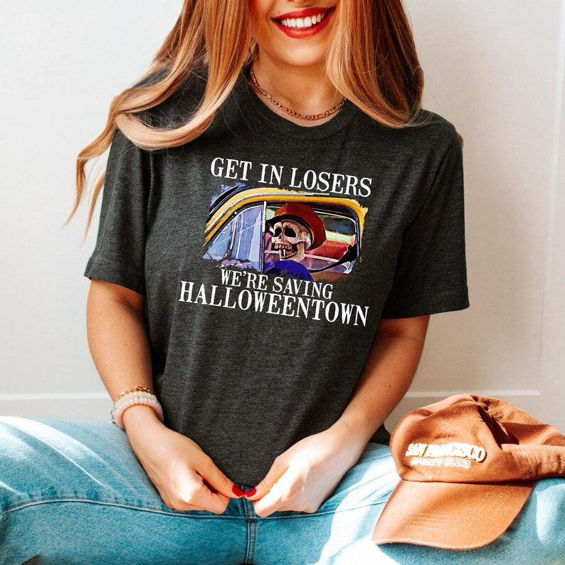 May include: A dark grey t-shirt with a graphic of a skeleton driving a yellow car. The text on the shirt reads "Get in losers, we're saving Halloweentown."