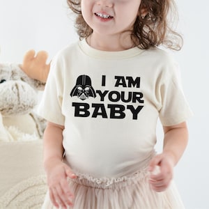 May include: A cream-colored t-shirt with the text "I AM YOUR BABY" in bold black letters, along with a black Darth Vader helmet graphic. The shirt is worn by a child.